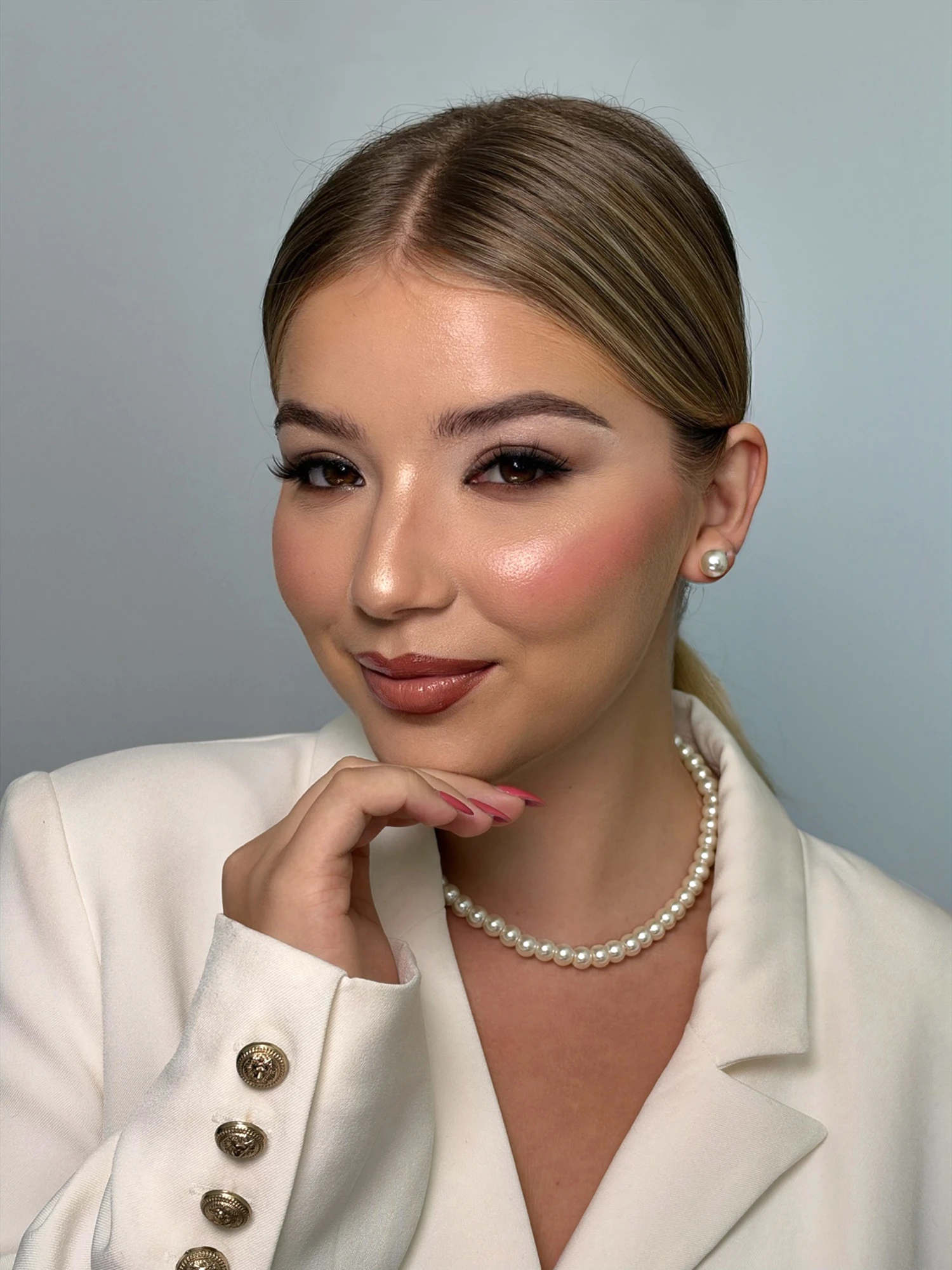 Smiling soft glam portrait with a sleek low ponytail, pearl studs and a nude glossy lip